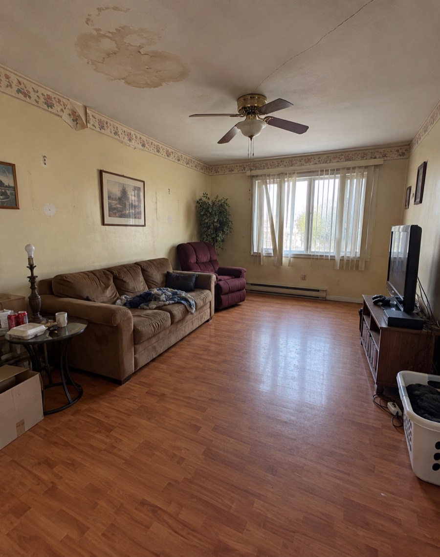 A tired living room: yellowed walls with a peeling floral wallpaper border, a brown water stain on the ceiling, a brass ceiling fan, a brown sofa, a burgundy recliner, broken vertical blinds and an orange laminate floor.