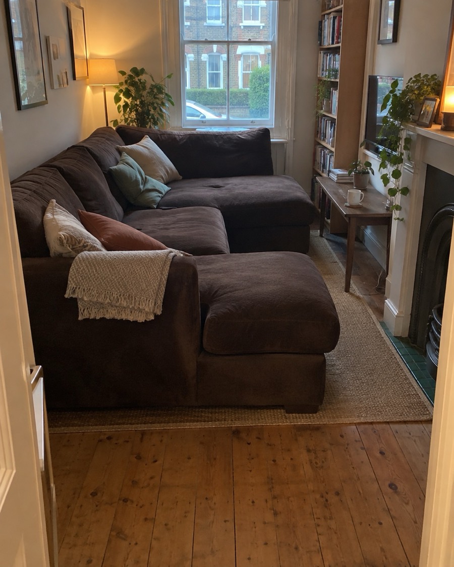 A dark brown velvet corner sofa with a long chaise filling a narrow living room almost up to the fireplace.